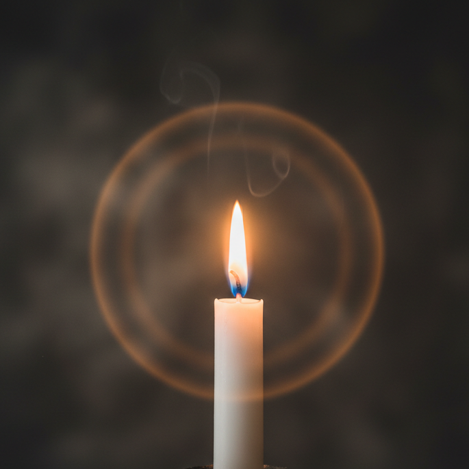 A single lit candle flame in sharp focus against a softly blurred dark background, with warm golden light radiating outward. Elegant, contemplative, and sophisticated mood. Minimal composition, fine art photography style.